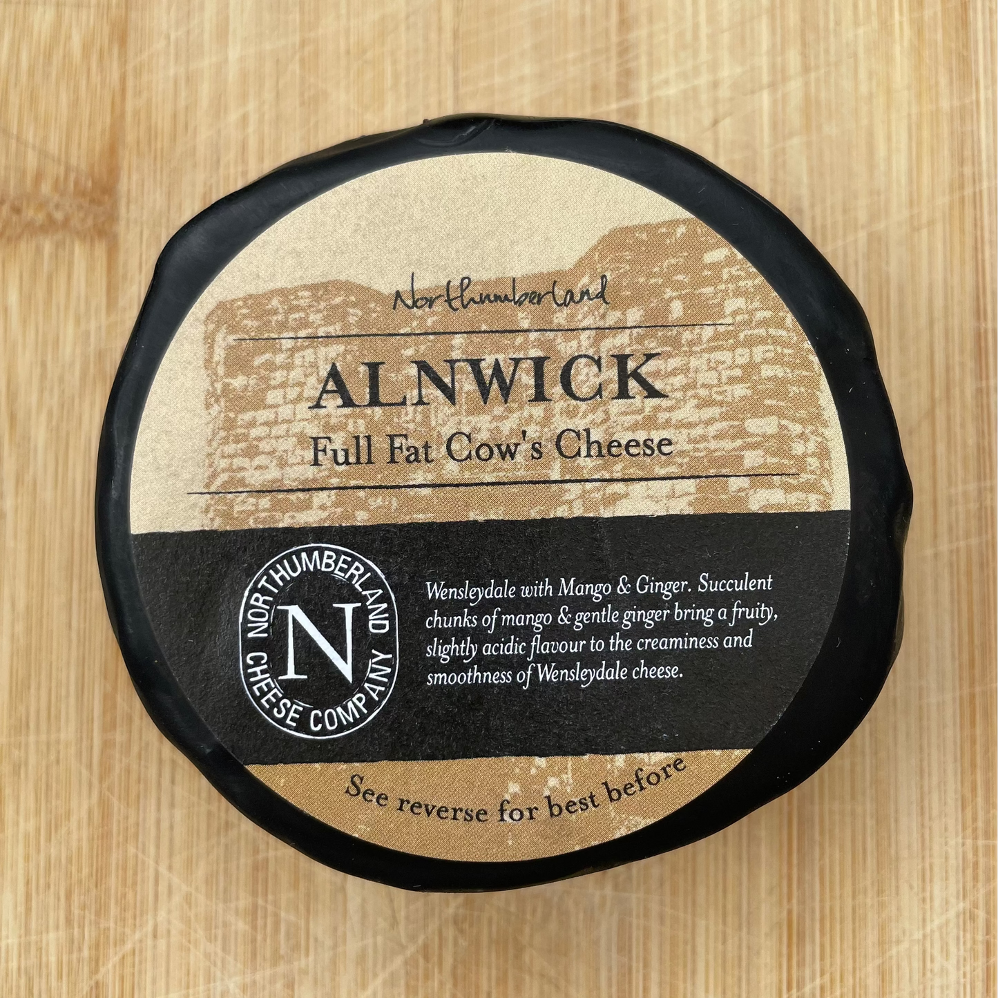 A round wheel of Alnwick Full Fat Cow's Cheese wrapped in black and kraft paper, placed on a light wood cutting board. The cheese wheel features a black and tan label with white text and a circular logo for 'Chester Northamptonshire Dairy'.