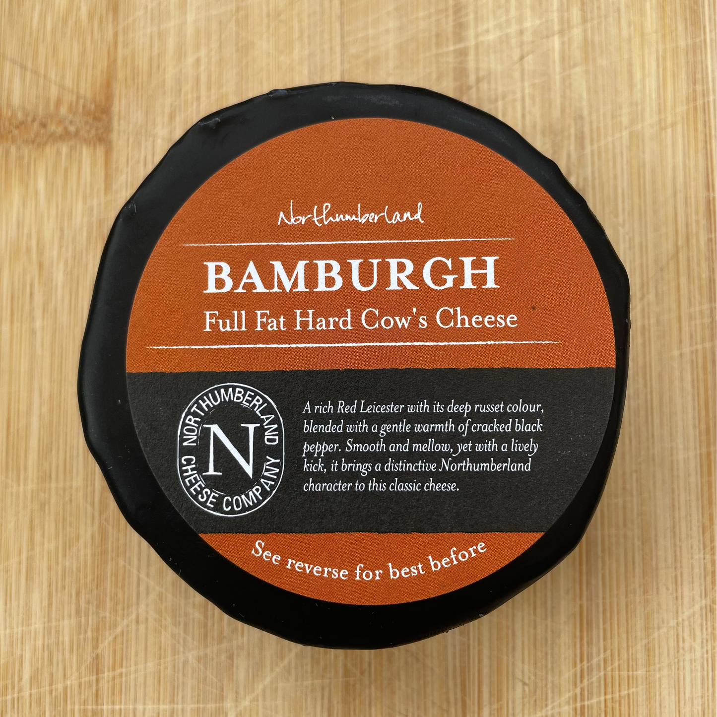 A round wheel of cheese wrapped in black wax and a brown paper label reading BAMBURGH, photographed from above on a light wood surface.