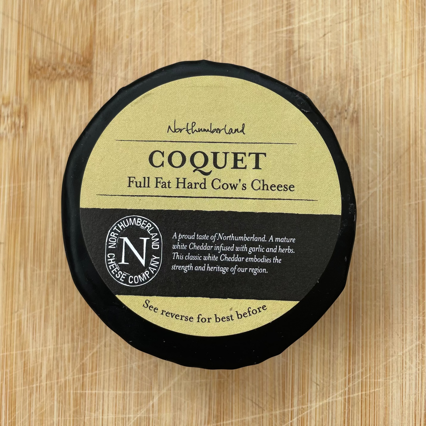 A round wheel of ZN Coquet Full Fat Hand Cow & Cheese cheese, wrapped in black paper with a tan and black label, placed on a light wood cutting board.