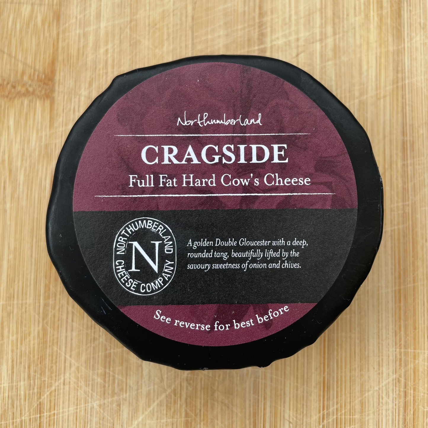 A round wheel of Cragside Full Fat Hard Cow's Cheese sits on a light wood cutting board. The cheese has a black plastic rim and a label split vertically into black and deep burgundy, with white text.