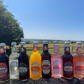 Eight bottles of Fentiman's fruit soda and ginger beer arranged in a row outdoors on a stone surface, with a green field and blue sky in the background. The bottles include Cherry Cola, Curiosity Cola, Gently Sparkling Elderflower, Mandarin Orange Jigger, Ginger Beer, Rose Lemonade, Dandelion & Burdock, and Raspberry Lemonade.