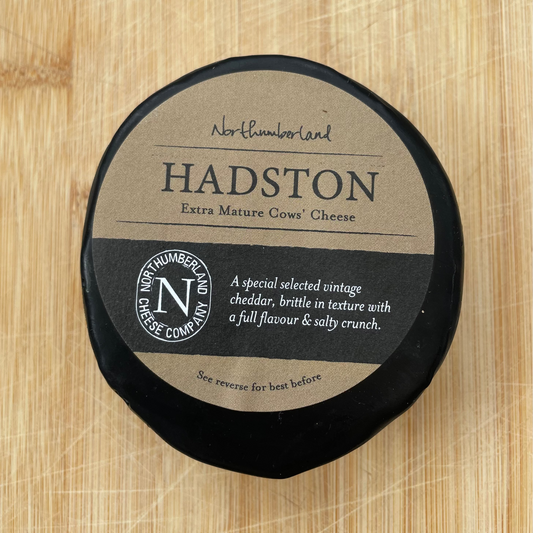 A round black cheese wheel with a kraft paper label reading 'HADSTON' placed on a light wood cutting board.